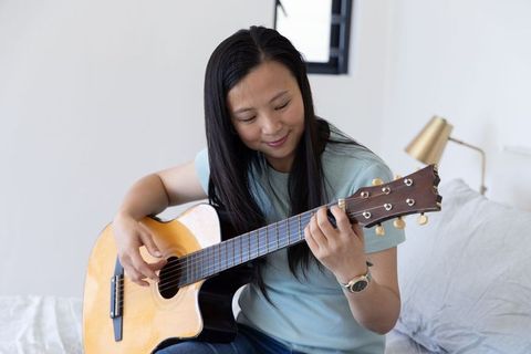 Asian woman practicing acoustic guitar on bed in calm minimalist bedroom interior