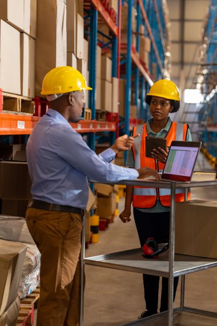 Foreman and female worker in warehouse discussing logistics using laptop and digital tablet. Both wearing safety helmets and vests, indicating focus on safety and efficiency. Ideal for illustrating teamwork, logistics, inventory management, and modern technology in industrial settings.