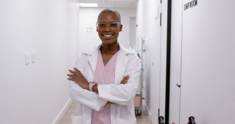 Confident african woman clinician standing in clinic hallway wearing white coat pink scrubs