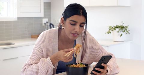 Woman Enjoying Noodles at Home While Checking Smartphone