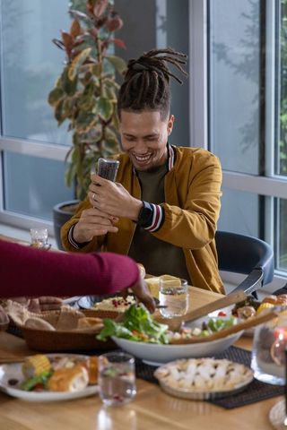 Smiling african american man seasoning salad at sunlit home dining table, convivial meal