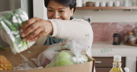 Senior indian woman unpacking groceries at sunlit kitchen counter pulling fresh greens