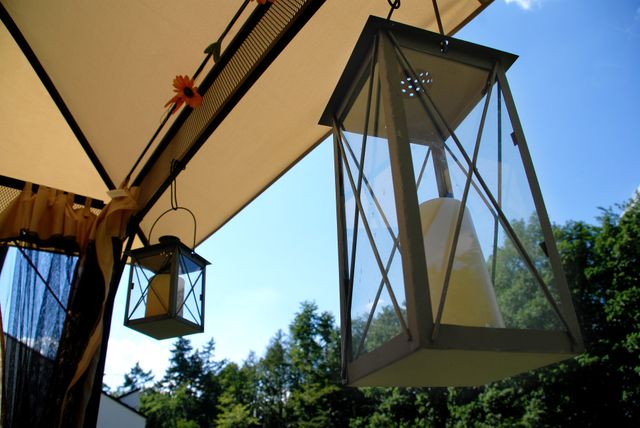 Lanterns hanging under a sunshade with trees and the sky in the background create a peaceful and rustic outdoor ambiance. Ideal for use in garden design inspiration, outdoor furniture catalogs, or advertisements promoting decorative items for patios and terraces.