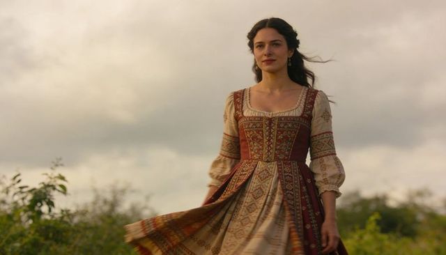 Woman walking in ornate renaissance dress through meadow under overcast sky windblown