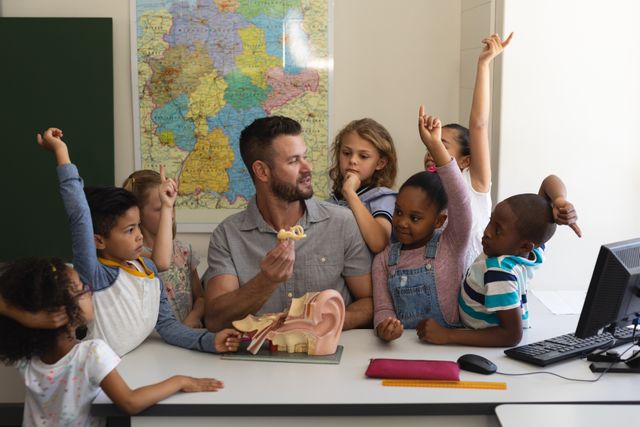 Front view of curious schoolkids raising hands around teacher teaching anatomy at desk in classroom of elementary school