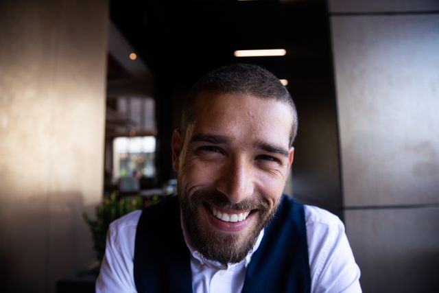 A close-up portrait of a happy, smiling, handsome Caucasian man with short dark hair and a beard, sitting inside a cafe. This image is perfect for use in lifestyle blogs, articles about digital nomads, advertisements for cafes or restaurants, and promotional materials for men's grooming products.