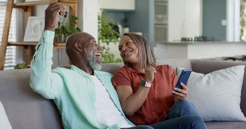 Mature diverse couple celebrating new home keys while relaxing on couch with smartphone