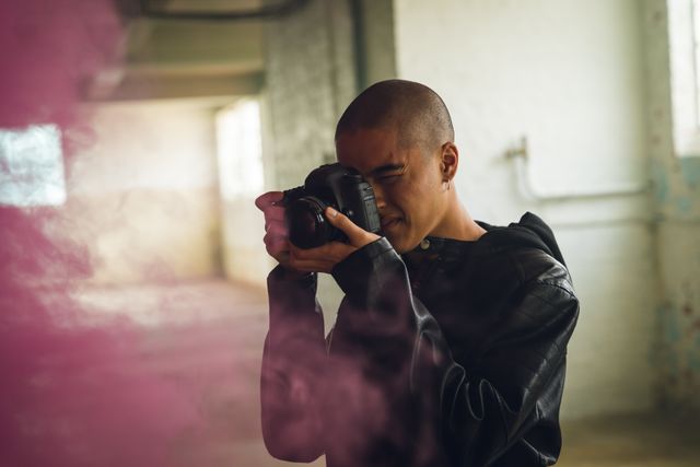 Young biracial man taking photos with an SLR camera in an empty warehouse, surrounded by pink smoke. He is wearing a black jacket, adding to the urban and creative atmosphere. Ideal for use in articles or advertisements related to photography, urban exploration, creativity, and artistic expression.