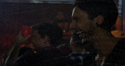 Laughing bearded man holding red cup at lively bar with crowd, candid nightlife moment