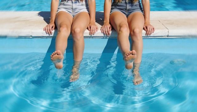 Two friends sitting on pool edge dipping bare feet in clear blue water sunny summer scene