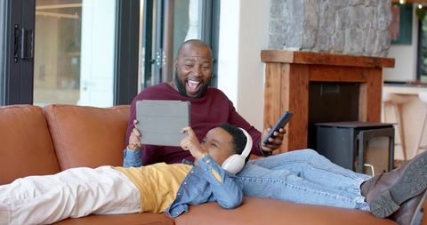 African american father and son relaxing on leather sofa sharing tablet and headphones