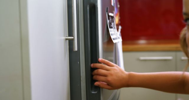 A child reaches out to open a refrigerator door, with copy space. Capturing a moment of independence, the image reflects a young one's curiosity and desire to explore.