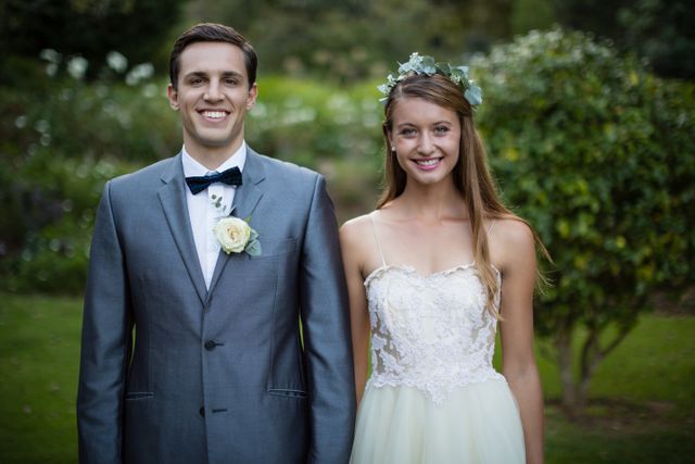 This image shows a smiling wedding couple standing together in a lush garden. The bride is wearing a white wedding dress with a floral crown, while the groom is dressed in a gray tuxedo with a bow tie and boutonniere. Ideal for use in wedding invitations, wedding planning websites, bridal magazines, and romantic greeting cards.