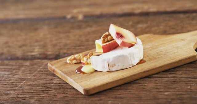 A slice of creamy brie cheese topped with a piece of fresh apple and a walnut drizzled with honey is presented on a wooden board. This appetizing setup suggests a sophisticated snack or a gourmet cheese platter element.