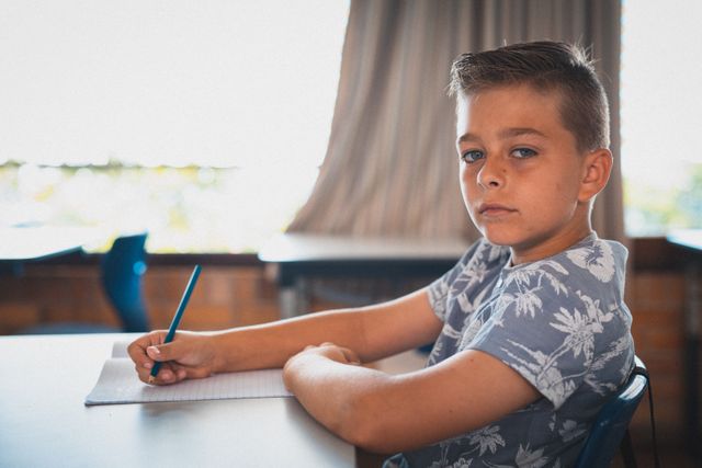 Caucasian schoolboy sitting at a desk in a classroom, writing in a notebook with a pencil. Ideal for educational content, school-related articles, and promotional materials for elementary schools. Can be used to illustrate themes of learning, concentration, and childhood education.