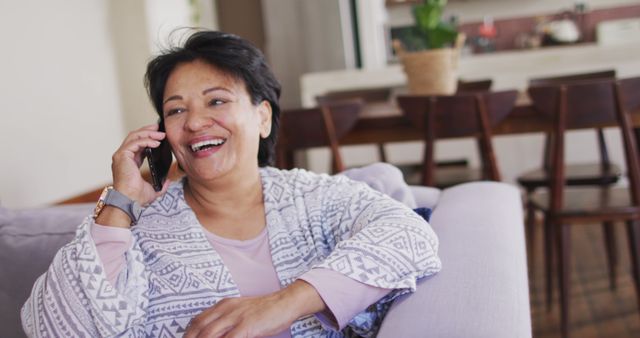Senior woman smiling and enjoying a phone call while sitting on a couch in a cozy living room. Ideal for use in communications, lifestyle, retirement, and family-related contexts. Perfect for promoting phone services, senior living, home comfort, and wellbeing advertisements.