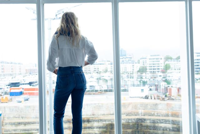 Young Caucasian businesswoman standing near a large window, looking out at an urban cityscape with buildings and waterfront. Ideal for use in business, professional, and workplace-related content, emphasizing modern office environments and thoughtful moments.