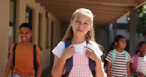 Sunlit multiracial students walking on school walkway with backpacks, girl in pink pinafore