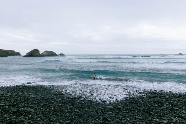 Surfer standing at the edge of the ocean on a pebble beach, facing large waves under a cloudy sky. Use in themes related to solitude, surf culture, nature, adventure, and outdoor recreation.