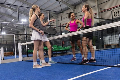Female padel players chatting at net on indoor blue court wearing sportswear and smiling