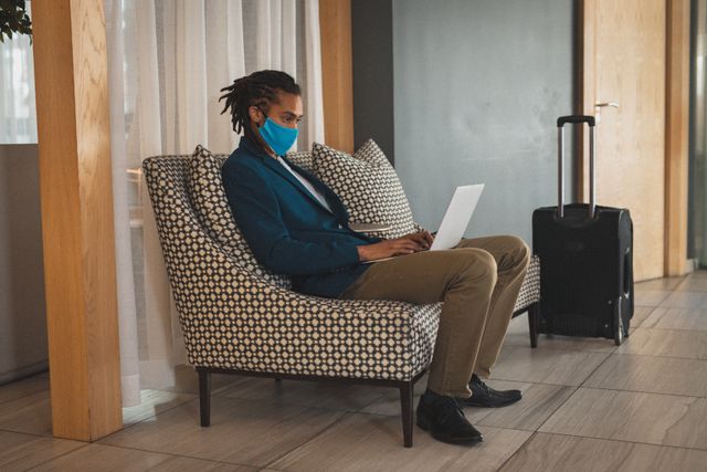 Biracial businessman wearing face mask sitting working in hotel lobby. business travel hotel during coronavirus covid 19 pandemic.