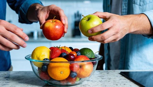 Two adults in denim shirts selecting apples from glass bowl of fresh fruit at kitchen counter