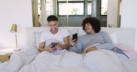 Diverse male friends laughing while using smartphones on bed