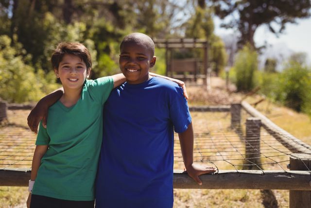 Two boys standing with arms around each other, smiling at an outdoor boot camp. Ideal for themes of friendship, teamwork, childhood adventures, and summer camp activities. Perfect for educational materials, youth programs, and advertisements promoting outdoor activities and bonding experiences.