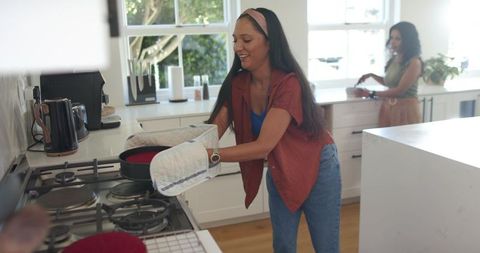 Diverse friends cooking together in sunlit modern kitchen removing hot skillet with mitts