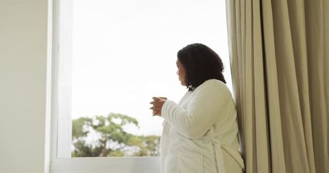African American woman gazing out window in white bathrobe holding mug morning calm