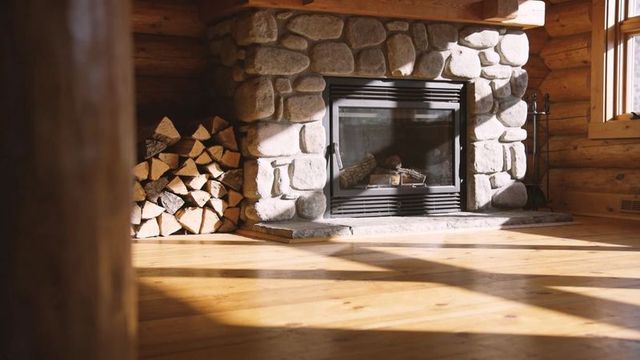 Sunlit stone fireplace in rustic log cabin with stacked firewood and toolset