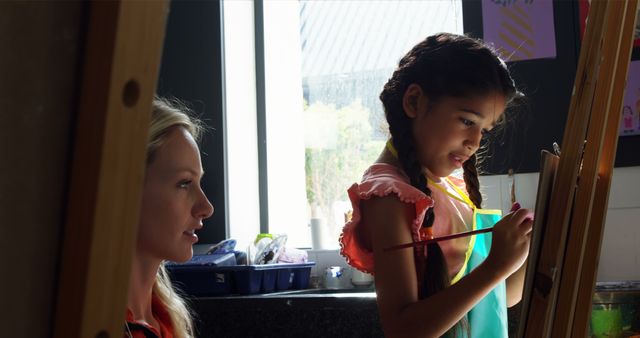 Young girl painting on easel with teacher providing guidance in art class. Great for educational materials, creativity workshops, school brochures, and promoting artistic learning environments.