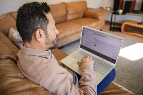 Asian man working on laptop on leather sofa cozy living room remote work productivity