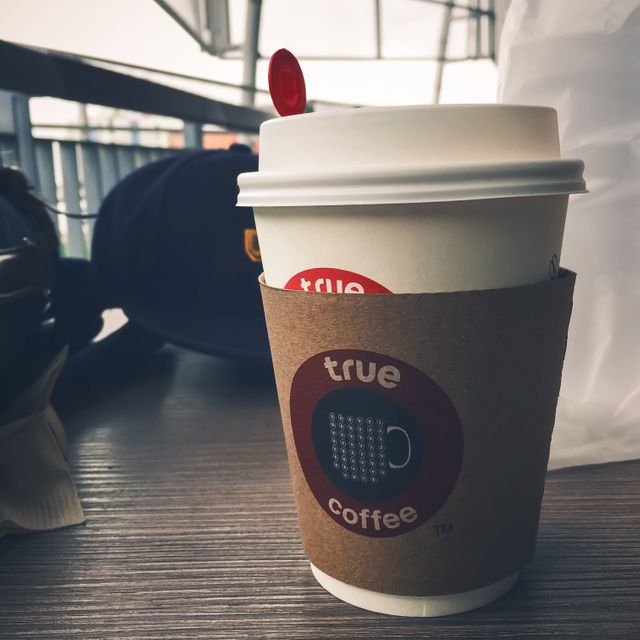 White disposable coffee cup with true coffee logo, cardboard sleeve, and red stirrer placed on wooden table. Urban cafe environment visible in the background, creating a relaxed and modern vibe. Ideal for lifestyle blogs, social media posts, branding materials, coffee-related content, advertisements, or campaigns focused on modern lifestyles and relaxation.
