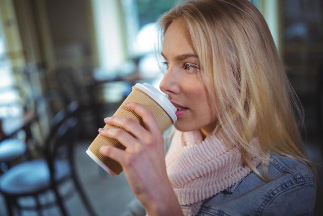 Blonde woman sipping coffee in a cozy cafe, wearing a pink scarf and denim jacket. Ideal for use in lifestyle blogs, coffee shop promotions, relaxation and leisure themes, and social media content related to casual dining and coffee culture.