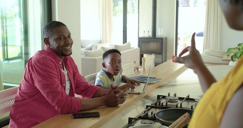 Smiling african american dad and son using laptop at bright open-plan kitchen island