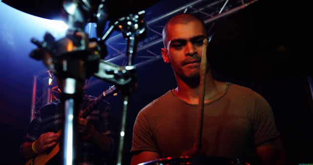 A young man is intently playing the drums on a stage, fully immersed in his performance during a live concert. The background shows stage lighting and musical equipment, enhancing the concert atmosphere. This can be used for event promotion, music industry content, concert advertisement, or articles related to live music performances.