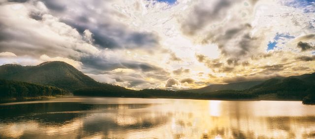 Golden sunset casting warm light over calm mountain lake with dramatic cloud formations. Perfect for nature blogs, travel brochures, meditation guides, or as a soothing wall art piece. Reflective water enhances tranquil atmosphere.