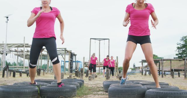 Happy caucasian female friends in pink t shirts crossing log bridge and tyres at bootcamp training. Female fitness, friendship, challenge and healthy lifestyle.