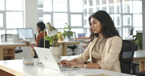 Indian businesswoman typing on laptop in modern open-plan office, teamwork and career focus