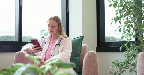 Female healthcare professional relaxing with smartphone and coffee in bright clinic lounge