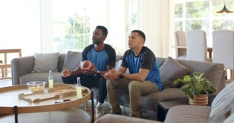 Diverse male friends watching football game on couch with pizza, chips and cold drinks