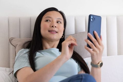Mature asian woman reclining on bed holding smartphone tapping screen calm lifestyle