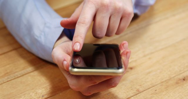 Close-up depicts a hand using a smartphone with one finger touching the screen against a wooden table background. Perfect for illustrating concepts related to technology usage, digital communication, mobile applications, and everyday tech habits. Ideal for marketing materials, tech blogs, and user experience design.