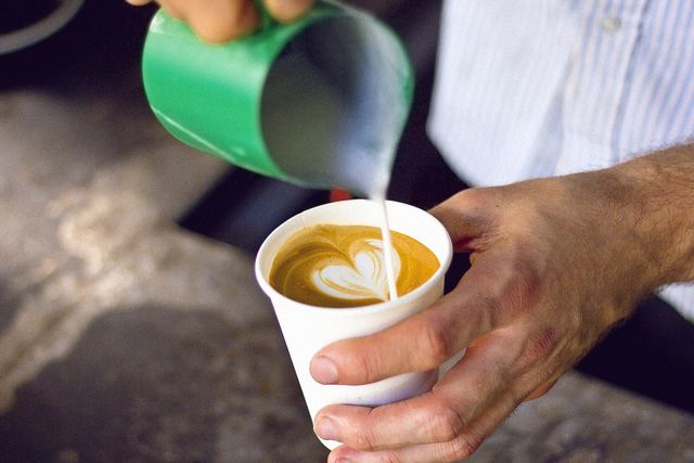 Barista pouring frothy milk into cappuccino with heart-shaped foam art in paper cup. Ideal for use in coffee shop promotions, barista training materials, and morning drink advertising. Could also be used for illustrating concepts related to artisanal beverages, caffeine culture, and hospitality services.