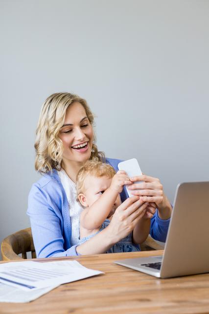Mother sitting with baby using smartphone at home workspace. Ideal for topics on parenting, multitasking, work-life balance, and home office setups. Could be used in articles related to technology use among young children, remote work environments, and family bonding time.