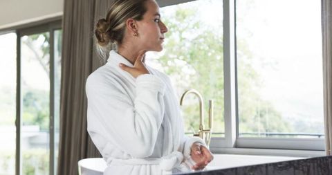 Luxury bathroom morning ritual woman in white bathrobe touching neck by window
