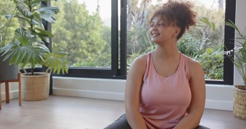 Young woman practicing mindfulness sitting crosslegged by window with plants and bright daylight