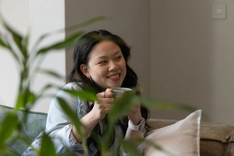 Asian woman enjoying coffee on sofa amid indoor plants smiling and looking sideways