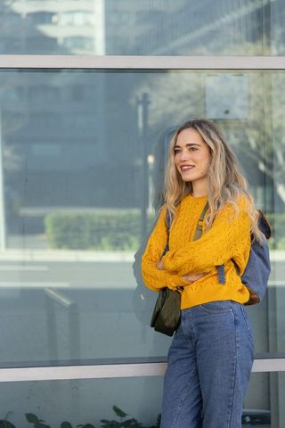 Young woman standing on sidewalk wearing mustard sweater and backpack, urban casual lifestyle